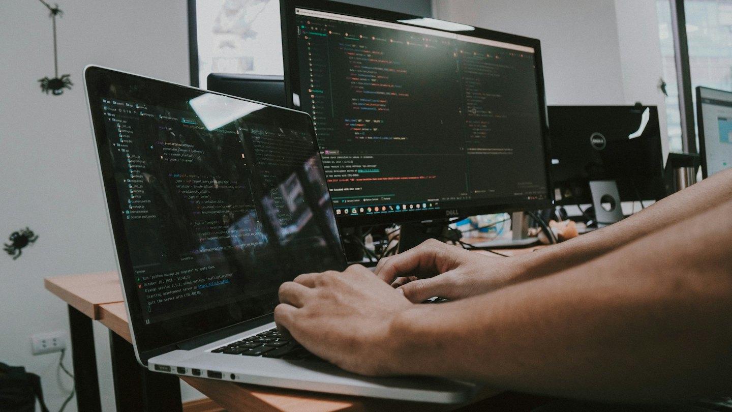 Hände tippen Code auf einem Laptop, der an einen großen Monitor angeschlossen ist.