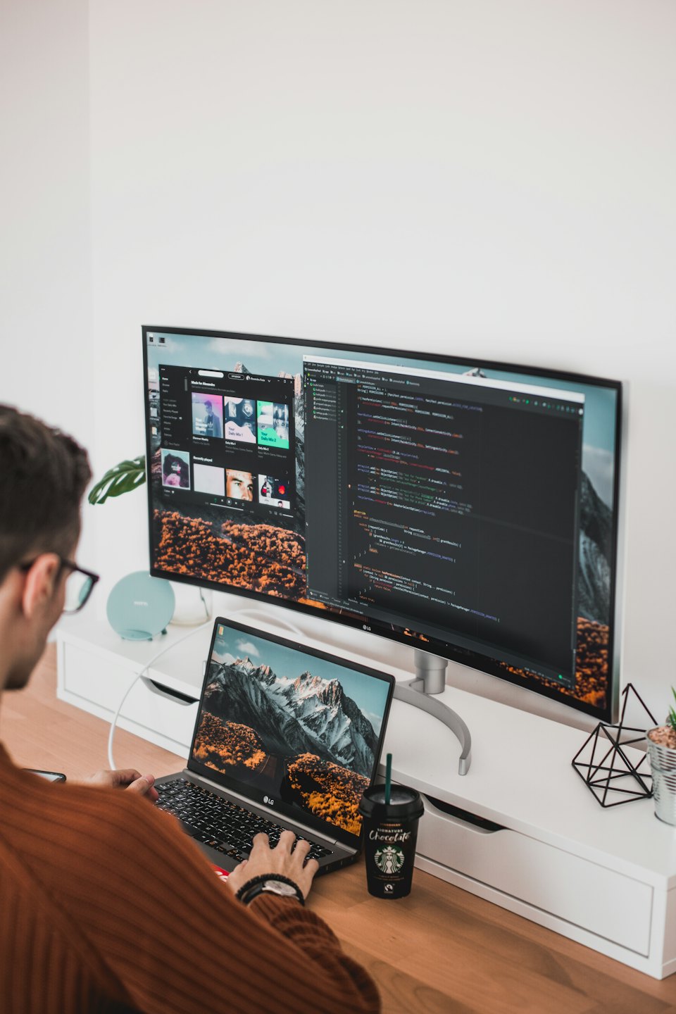Eine Person mit Brille arbeitet am Schreibtisch mit Laptop und Monitor.